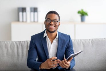 Portrait of happy black male psychologist looking at camera and taking notes during therapy session at clinic. Friendly millennial African American psychotherapist posing and smiling at office